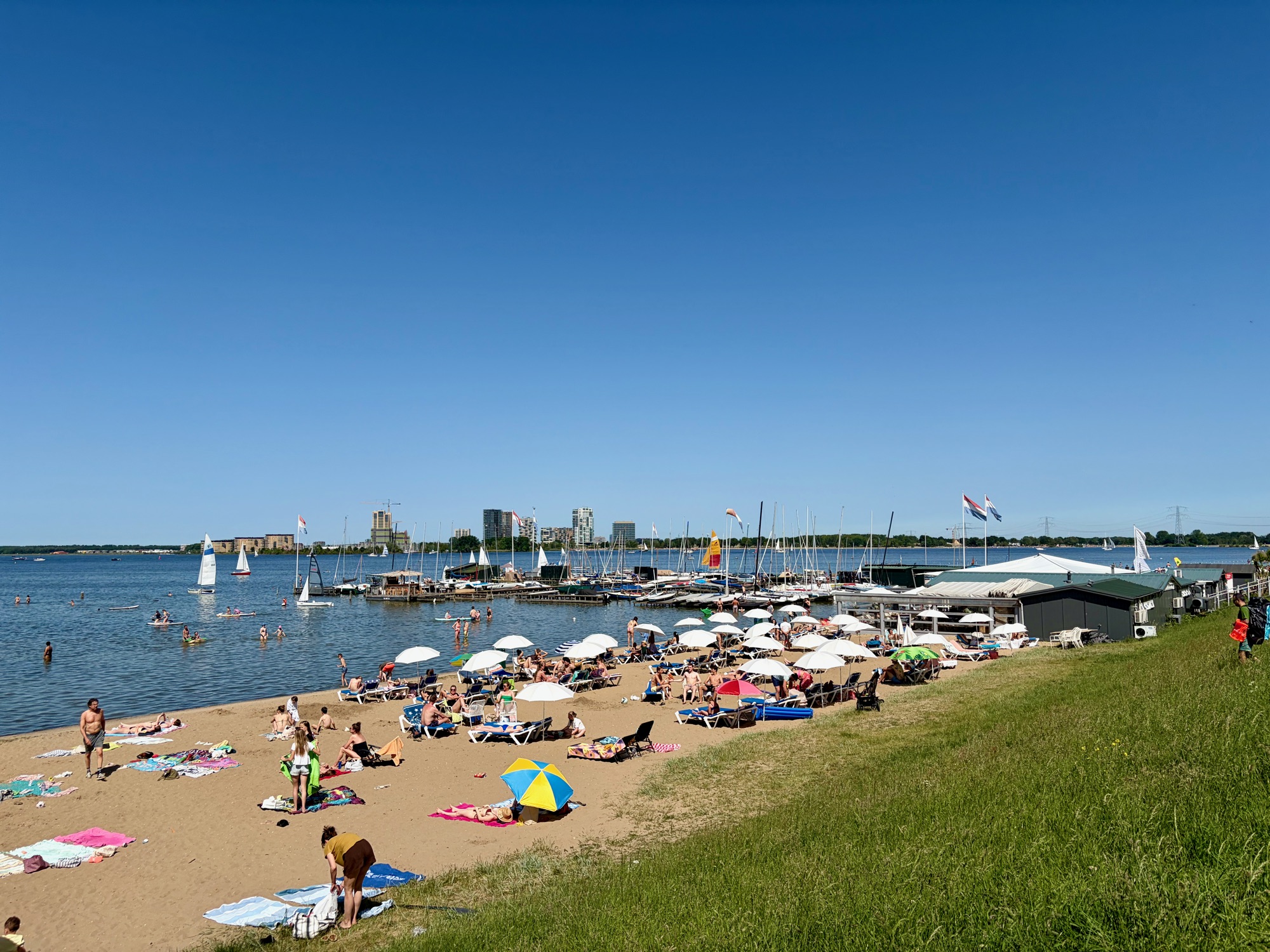 Strand Muiderberg vanaf het dijktalud: parasols, ligbedden, zwemmers, zeilboten in de jachthaven en de skyline van IJburg aan de overkant.